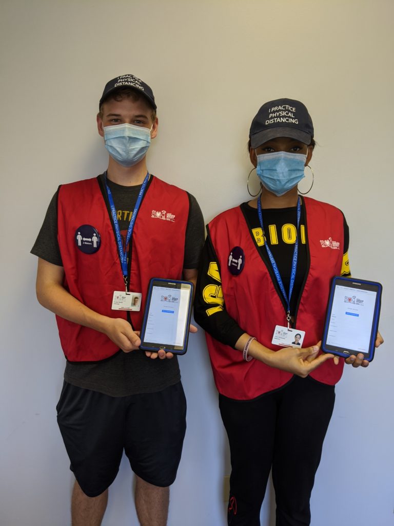 Two people wearing masks, name badges, and OHS vests holding iPads